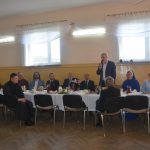 Grupa elegancko ubranych osób zasiada przy bogato zastawionym stole na sali. Męzczyzna zasiada za pomocą mikrofonu.