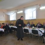 Grupa elegancko ubranych osób zasiada przy bogato zastawionym stole na sali. Przed stołem mężczyzna ubrany w sułtanne.