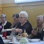 Grupa osób róznej płci i wieku zasiadajaca przy stołach na sali.