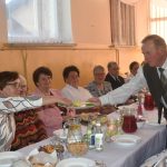 Duża grupa osób róznej płci i wieku zasiadajaca przy stołach na sali. Jeden z mężczyzn podaje kawałki ugotowanych jajek biesiadnikom.