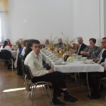 Duża grupa osób róznej płci i wieku zasiadajaca przy stołach na sali.