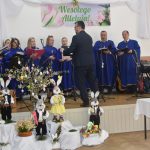 Chór kobiet i mężczyzn w granatowych strojach i męzczyzna w czarnym garniturze na scenie.