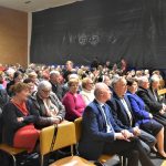 Duża grupa osób różnej płci i wieku siedząca na na sali gimastycznej.