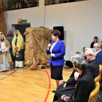 Starsza kobieta przemawijąca za pomocą mikrofonu na sali gimastycznej. W tle aktorzy w strojach nowotestamentalnych oraz widzowie.