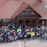 Duża grupa dorosłych i dzieci pozująca do zdjecia przed wielkim, drewnianym budynkiem.