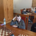 Chłopiec siedzący przy stoiku z szachami. W tle grupa dzieci grająca w szachy.