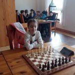 Dziewczynka siedząca przy stoiku z szachami. W tle grupa dzieci grająca w szachy.