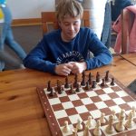 Chłopiec siedzący przy stoiku z szachami. W tle grupa dzieci grająca w szachy.