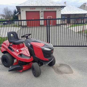 Czerwono-czarny traktorek ogrodowy na tle budynku OSP.