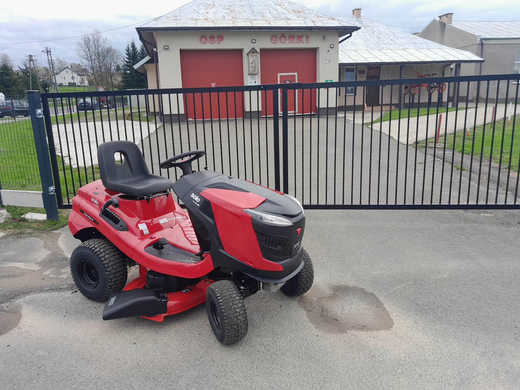 Czerwono-czarny traktorek ogrodowy na tle budynku OSP.