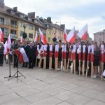 Duża grupa dorosłych i młodzieży z polskimi flagami stojący na rynku.