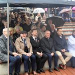 Grupa osób siedząca na schodkach na otwartej przestrzeni. Pada deszcz a ludzie zaslaniaja się parasolami i namiotami.