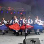 Grupa tancerzy w stojach ludowych tańczy na scenie na wolnym powietrzu..