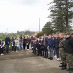 Grupa młodzieży stojąca na placu. W tle drzewa i smaochody.