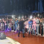 Duża grupa osób za barierkami obserwująca występ muzyczny w nocy.