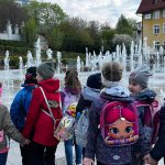 Grupa dzieci przedszkolnych wraz z opiekunami podczas przechadzki po mieście. Na pierwszym tle fontanna.