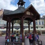 Grupa dzieci przedszkolnych wraz z opiekunami podczas przechadzki po mieście i odpoczynku przy zabytkowej studni.