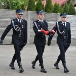 Trzech paradujących strażaków. Środkowy niesie w rękach polską flagę