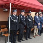 Grupa strażaków i gości w garniturach stojąca pod namiotem.