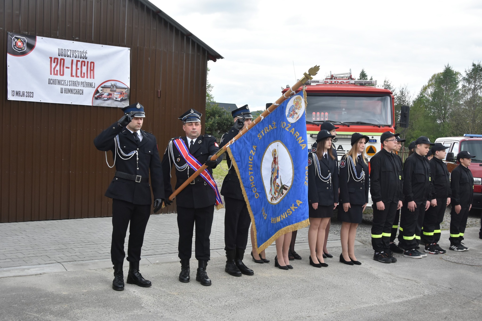 Grupa strażaków z pocztem sztandarowym na uroczystej zbiórce.