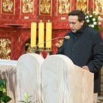 Mężczyzna w ciemnym płaszczu przemawia do mikrofonu przy mównicy w kościele.