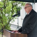 Starszy mężczyzna w garniturze przemawia za pomoca mikrofonu przed dużym budynkiem.