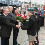 Trzech mężczyzn przekazujących kwiaty i znicze harcerzom. W tle orkiestry i poczty sztandarowe.