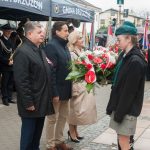 Dwóch mężczyzn i kobieta przekazujący kwiaty i znicze harcerzom. W tle orkiestry i poczty sztandarowe.