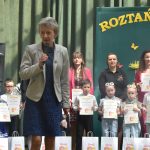 Starsza kobieta przemawia za pomoca mikrofonu. W tle grupa dorosłych i dzieci z dyplomami.