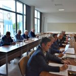 Grupa osób siedząca przy stolikach na sali konferencyjnej. Na stołach znajdują się butelki wody, szklanki i dokumenty.