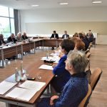 Grupa osób siedząca przy stolikach na sali konferencyjnej. Na stołach znajdują się butelki wody, szklanki i dokumenty.