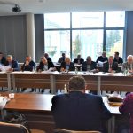 Grupa osób siedząca przy stolikach na sali konferencyjnej. Na stołach znajdują się butelki wody, szklanki i dokumenty.