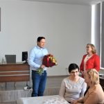 Męzczyzna z bukietami kwiatów stojący na sali konferencyjnej. Obok stoi starsza kobieta i trzy panie siedzą przy pobliskim stoliku.