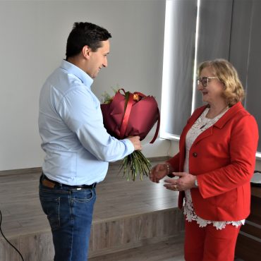 Mężczyzna w jasnej koszuli przekazuje kwiaty starszej kobiecie w czerwonej marynarece.