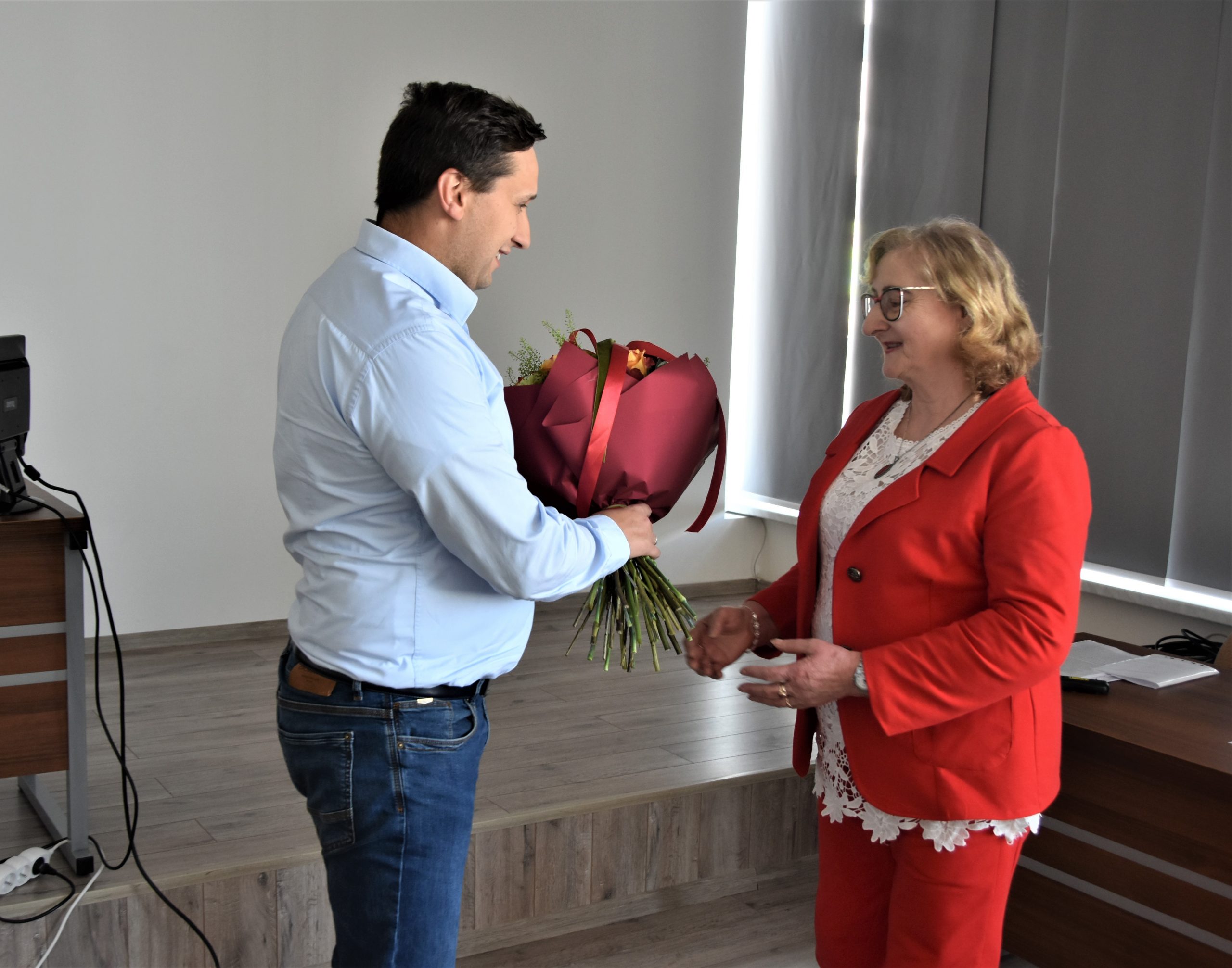 Mężczyzna w jasnej koszuli przekazuje kwiaty starszej kobiecie w czerwonej marynarece.