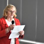 Starsza pani przemawia na sali konferencyjnej za pomocą mikrofonu,. W ręce trzyma kartkę papieru.