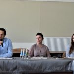 Mężczyzna w błękitnej koszuli oraz dwie kobiety siedzący przy podłużnym stole.