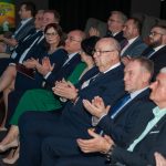 Grupa osób dorosłych osób w odswiętnych strojach siędzacych na slai kinowej i klaszczących.