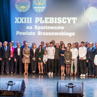 Grupa osób w odświetnych strojach pozująca do zdjecia przed ekranem kinowym.