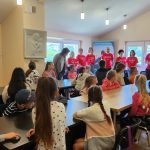 Grupa dzieci siedzi przy stole w tle stoją dorośli w czerwonych koszulkach