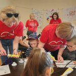 Dzieci siedzą przy stole, 4 kobiety pomagają dzieciom w pracach