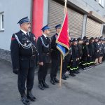Grupa ludzi w mundurach strażackich, jeden z mężczyzn trzyma sztandar