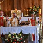 Trzech duchownych w szatach stoją przy ołtarzu i trzymają ręce podniesione do góry.