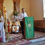 Mężczyzna przy mównicy w kościele, w tle siedzi dwóch duchownych w szatach.