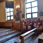 Mężczyzna przy mównicy w kościele. Obok stoją kobiety w strojach ludowych.