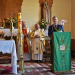 Mężczyzna przy mównicy w kościele, w tle stoi dwóch duchownych w szatach.