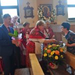 Mężczyzna stoi z kwiatkiem, w tle kobiety w strojach ludowych. Jedna z nich podaje drugiej dłoń.