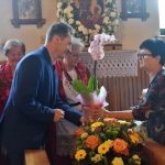 Mężczyzna wręcza kobiecie kwiaty, w tle stoją kobiety w strojach ludowych