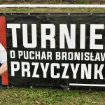 Baner czarny z napisami białymi i fotografią mężczyzny w białej koszuli, baner wisi na metalowym ogrodzeniu.