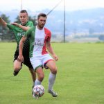 Mężczzni jasnej karnacji ubrani w strje sportowe grają w piłkę nożną na boisku.